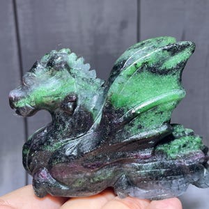 May include: A carved stone dragon figurine featuring a vibrant mix of green, black, and deep magenta mineral inclusions. The sculpture displays a detailed head with a crest, prominent wings folded against its body, and a textured, scaled surface.