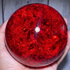 Puede incluir: Un objeto esférico con una superficie exterior lisa y transparente, y un interior de color rojo intenso y vibrante con una textura cristalina fracturada. La estructura interna crea una apariencia compleja y facetada que refleja la luz.