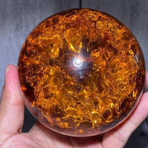 May include: A spherical object made of clear, polished resin or glass, featuring a dense, fractured interior with vibrant amber and gold-toned metallic foil flakes that create a reflective, multi-faceted appearance throughout the sphere.