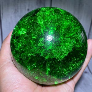 May include: A spherical object made of clear material containing a vibrant green, fractured, crystalline-like interior. The surface is smooth and polished, reflecting light with a bright white circular highlight near the centre of the sphere.