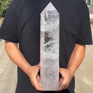 May include: A large, clear quartz crystal point with a hexagonal shape and a pointed top. The crystal features natural internal inclusions, fractures, and cloudy white patches throughout its transparent, colorless body, showcasing a polished finish.