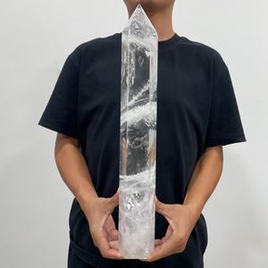 Op de afbeelding: Een grote, heldere, zeshoekige kwartskristallen punt met een spitse bovenkant. Het kristal bevat interne witte insluitingen en breuken die een wervelend, troebel patroon vormen door de transparante, kleurloze en langwerpige minerale structuur.