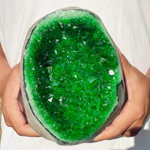 Könnte beinhalten: Eine große, oval geformte Geode mit einem tiefen, leuchtend smaragdgrünen kristallinen Inneren. Die äußere Schale des Steins ist in einem neutralen, matten Grau gehalten, das einen starken Kontrast zu den facettierten, grünen Kristallen bildet.