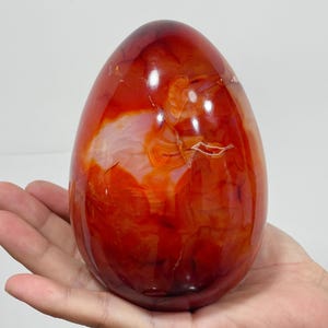 Può includere: Una scultura in pietra lucida a forma di uovo caratterizzata da una vibrante tavolozza di colori arancione intenso e rosso traslucido, con bande interne naturali, motivi vorticosi e sottili inclusioni bianche su tutta la superficie liscia e lucida.