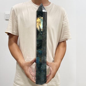 Könnte beinhalten: Ein hoher, polierter Labradorit-Kristallturm in Form eines hexagonalen Prismas. Der Stein hat eine dunkelgrüne und anthrazitfarbene Grundfarbe mit einem markanten, irisierenden goldgelben Schimmer im oberen Bereich der facettierten Oberfläche.