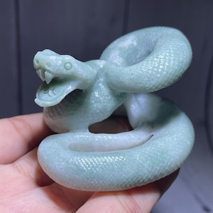 May include: A pale green carved stone figurine of a coiled snake with its mouth open, revealing fangs. The sculpture features a textured, scale-like surface pattern across its body and a smooth, polished finish on the head and interior coils.