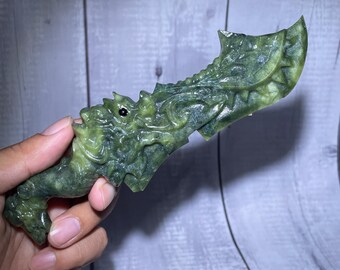 May include: A carved green stone dagger featuring an intricate dragon head design on the handle and a curved, textured blade. The stone displays natural variations in deep forest green and lighter olive tones with a polished, smooth finish.