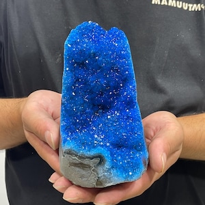 May include: A vibrant blue crystal geode specimen featuring a dense cluster of sparkling, deep azure mineral formations. The geode has a rough, grey, natural stone exterior base that supports the upright, jagged, and crystalline blue interior structure.