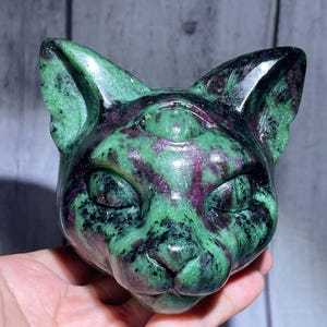 May include: A polished cat head carving crafted from green zoisite with deep red ruby inclusions. The sculpture features pointed ears, closed eyes, and a small third eye detail centered on the forehead, showcasing a smooth, glossy stone texture.
