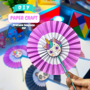 Puede incluir: Un abanico de papel circular con un diseño plisado en morado y blanco, con un recorte central de una cabeza de unicornio de dibujos animados. El unicornio tiene un cuerno amarillo, pelo morado y turquesa, y grandes ojos morados. Una varilla turquesa está unida.