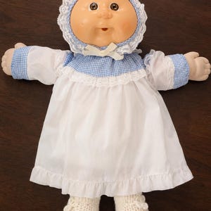 May include: A vintage doll with a round face, wearing a white dress with a ruffled hem and a blue gingham collar and cuffs. The doll has a white lace bonnet and matching knitted booties. The doll's skin is a peach colour, and it has brown eyes.