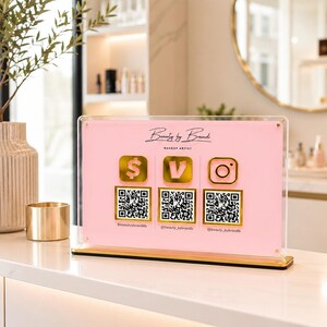 May include: A pink acrylic sign with gold accents displays the text "Beauty by Brandi, Makeup Artist." It features three gold icons: a dollar sign, a "V," and an Instagram logo, each with a corresponding QR code and social media handle.