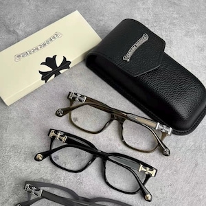 Peut inclure: Lunettes avec des montures noires, marron et grises, ornées de détails en forme de croix. Un étui en cuir noir et une boîte crème avec le texte "CHROME HEARTS" sont également présents. Les lunettes sont de forme carrée.
