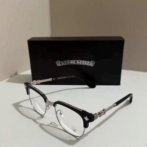 Puede incluir: Gafas negras y plateadas con un diseño de montura semi al aire y detalles metálicos en las varillas y el puente. Las lentes son transparentes y el estuche rectangular negro al fondo muestra el logotipo de Chrome Hearts en letras blancas.