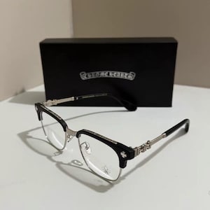 Puede incluir: Gafas negras y plateadas con montura rectangular y diseño de cruz decorativa en las patillas. Las gafas están colocadas delante de una caja negra con un logotipo plateado. Las lentes son transparentes.