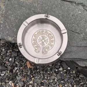 May include: A circular silver-toned metal ashtray featuring a brushed finish. The center is engraved with a circular logo containing the text Chrome Hearts and a central cross emblem. Four small cross motifs are embossed on the outer rim.