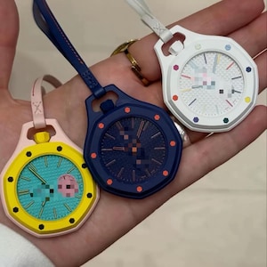 May include: Three octagonal pocket watches with attached straps. One is yellow with a teal face, one is dark blue with a matching face, and one is white with a white face featuring colorful hour markers. Each watch has a textured dial pattern.