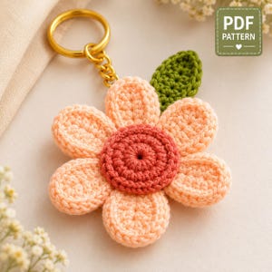 May include: A crochet flower keychain featuring a dark coral center, six light peach petals, and a single green leaf. The flower is attached to a gold-toned metal split ring. A green rectangular badge in the corner displays the text PDF PATTERN.