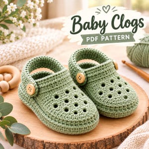 Könnte beinhalten: Ein Paar salbeigrüne gehäkelte Babyschuhe in Clog-Optik mit perforiertem Zehenbereich und einem beweglichen Fersenriemen, der mit einem hellen Holzknopf befestigt ist. Der Text Baby Clogs PDF PATTERN steht in dunkelgrüner Schrift darüber.