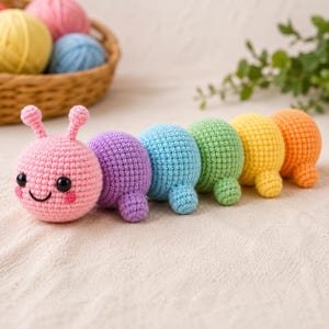 May include: A multi-colored crochet caterpillar toy featuring a pink head with antennae, black eyes, and a stitched smile, followed by segments in purple, blue, green, yellow, and orange. Each round segment has a small nub for a foot.