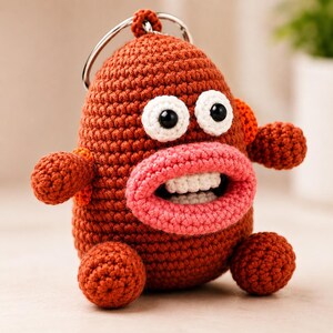 May include: A brown crochet keychain shaped like a creature with a large pink mouth, white teeth, and two round white eyes with black pupils. The figure has small round arms and legs, and a silver metal keyring attached to the top of its head.
