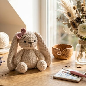 May include: A crocheted bunny with a pink bow sits on a wooden surface, next to a ball of yarn, a small bouquet of lavender, and a mug. A vase of flowers and a wooden bowl are also on the table. The bunny is beige.