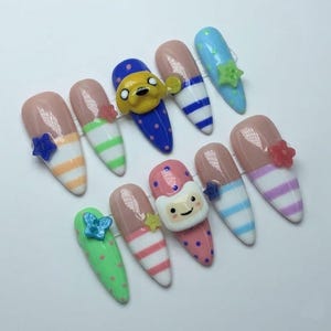 May include: A set of ten almond-shaped press-on nails featuring various designs including horizontal stripes, polka dots, and solid colours. Several nails are adorned with small 3D charms shaped like stars, flowers, and cartoon character faces.