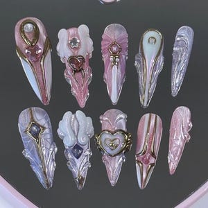 May include: A set of ten press-on nails featuring almond and stiletto shapes in shades of pink, white, and iridescent purple. The designs include 3D heart charms, gold metallic accents, white sculpted wings, and embedded crystals on a glossy finish.