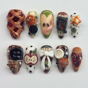 May include: A set of ten press-on nails featuring autumn-themed 3D decorations including a lattice pie, pumpkins, an apple with a worm, music notes, stars, black buttons, a white bow, and plaid patterns in shades of brown, green, white, and black.