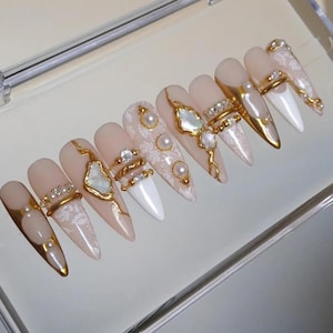 May include: A set of ten stiletto-shaped press-on nails featuring a nude and white color palette with intricate lace patterns, gold metallic accents, embedded white pearls, and clear rhinestones arranged in various decorative designs.