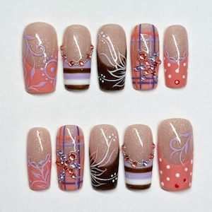 May include: A set of ten press-on nails featuring a nude glitter base with various designs including coral floral patterns, brown and white horizontal stripes with pink rhinestones, white floral art, plaid patterns with purple gems, and coral polka dots.