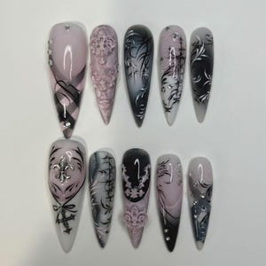 May include: A set of ten stiletto-shaped press-on nails featuring a color palette of soft pink, black, and white. The designs include intricate black filigree patterns, raised 3D baroque-style ornaments, silver fleur-de-lis charms, and small clear rhinestones.