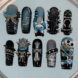 May include: A set of ten press-on nails featuring a black and blue color palette with various 3D embellishments including silver metal charms, chains, spikes, a cat figure, a skull, a cross, a bow, and star accents with zebra and lace patterns.