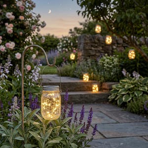 May include: A glass jar lantern filled with fairy lights hangs from a metal hook in a garden. Other glowing lanterns are placed along a stone pathway and steps. The scene is surrounded by greenery, flowers, and a crescent moon in the sky.