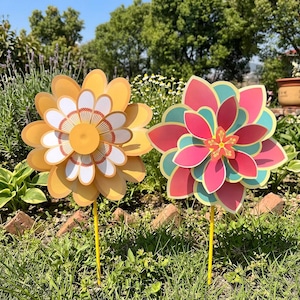 May include: Two flower-shaped pinwheels on yellow sticks. The left pinwheel features layered yellow and white petals with a central orange circle. The right pinwheel displays layered pink and teal petals with a small orange star-shaped centre.