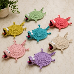 Op de afbeelding: Een verzameling van zeven gehaakte dierenfiguren in verschillende kleuren, waaronder groen, geel, roze, teal, rood, beige en paars. Elk figuur heeft een rond lichaam, vier kleine pootjes, een staart, een wijd open mond en twee witte ogen.