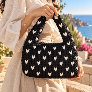 Könnte beinhalten: Eine schwarze Häkeltasche mit einem sich wiederholenden Muster aus kleinen weißen Herzformen. Die Tasche hat eine strukturierte, abgerundet rechteckige Form mit einem dicken, integrierten Griff an der Oberseite aus grobem Garn.