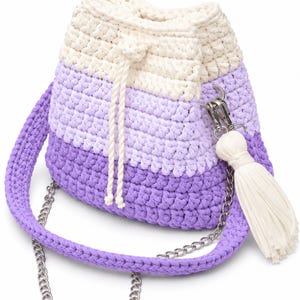 May include: A crochet bucket bag featuring a color-blocked design in cream, light purple, and dark purple. The bag includes a cream drawstring closure, a matching purple crochet shoulder strap, a silver-tone metal chain, and a cream tassel.