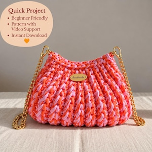May include: A crochet handbag featuring a ribbed texture in alternating shades of bright orange and light pink. The bag includes a gold-tone metal chain strap attached with lobster clasps and a small oval gold-tone metal tag labeled handmade.