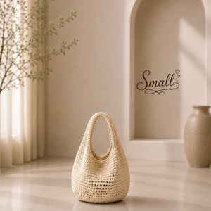 Puede incluir: Un bolso de estilo hobo tejido a ganchillo en color crema, con un asa integrada &uacute;nica y una base redondeada. El bolso presenta un patr&oacute;n de puntos denso y texturizado. La palabra Small est&aacute; escrita en cursiva marr&oacute;n oscuro sobre el bolso.