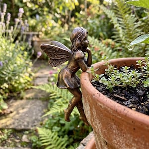 Puede incluir: Una figura de hada de color bronce con alas detalladas y una pose pensativa, posada en el borde de una maceta de terracota. El hada lleva un vestido corto y botas. El fondo presenta un exuberante jardín con follaje verde y flores moradas.