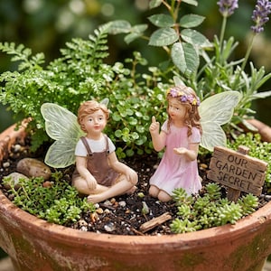 Puede incluir: Dos figuras de hadas con alas translúcidas sentadas en una maceta de terracota llena de vegetación. Una hada viste un mono marrón, la otra un vestido rosa y una corona de flores. Un pequeño letrero dice "OUR GARDEN SECRET."