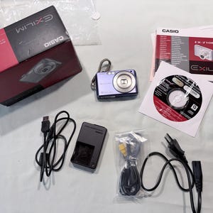 May include: A purple Casio Exilim digital camera with a wrist strap, accompanied by its original black and red box, a user manual, a software CD, a battery charger, a USB cable, and a power cord with a plug, all displayed as a complete set.