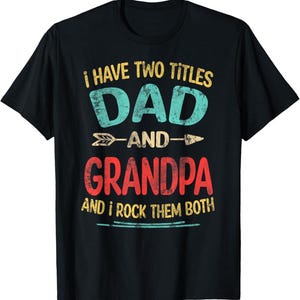 Pode incluir: Camiseta preta com a frase "I HAVE TWO TITLES DAD AND GRANDPA AND I ROCK THEM BOTH" em estilo vintage. "DAD" é turquesa e "GRANDPA" é vermelho. Inclui setas decorativas.