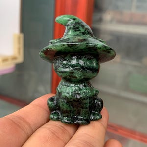 May include: A small, carved stone figurine of a frog wearing a pointed witch hat. The sculpture is made of a dark green mineral with black mottled patterns throughout, featuring large, round, carved eyes and a textured surface on the hat and body.