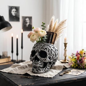 May include: A decorative skull-shaped vase in silver and black, filled with flowers and pampas grass, sits on a dark wooden table. The vase has intricate carved details. Other items include candles, books, and vintage portraits.