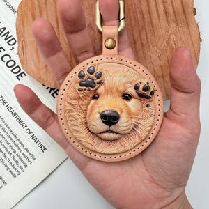 May include: A round, tan leather keyring featuring a detailed, embossed, and hand-painted golden retriever puppy face with two raised paw prints. The keyring is attached to a brass-coloured metal lobster clasp with a matching rivet.