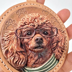 May include: A circular tan leather keyring featuring a detailed embossed portrait of a brown poodle wearing dark-rimmed glasses and a green and white striped scarf. The background includes embossed paw prints and hearts with white stitching.