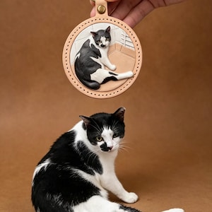 May include: A round, tan leather keyring featuring a raised, hand-painted relief of a black and white cat sitting on a tiled floor. The leather disc is stitched along the edge and attached to a gold-toned metal carabiner clip at the top.