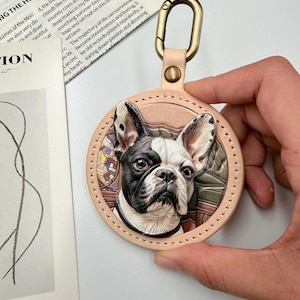 May include: A circular tan leather keyring featuring a three-dimensional embossed portrait of a black and white French Bulldog. The dog is centred within a stitched leather border, attached to a brass-toned metal lobster clasp hardware.